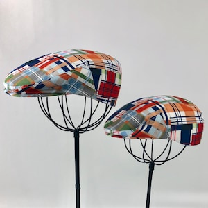 May include: Two patterned newsboy caps on display stands. The caps are made of a patchwork fabric with red, blue, green, and white squares.