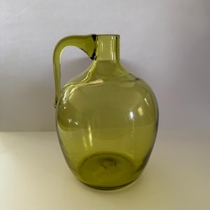 Puede incluir: Una jarra de vidrio verde con asa. La jarra es transparente y tiene un cuerpo redondo.