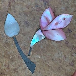 Puede incluir: Primer plano de una flor hecha de piezas de concha iridiscente, con una gama de colores rosa y azul, incrustada en una superficie de piedra marrón.