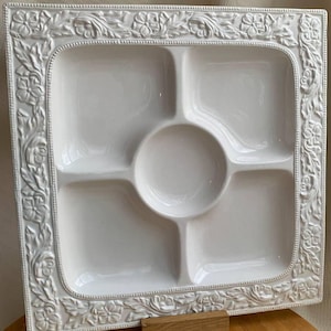Op de afbeelding: Een witte vierkante serveerschaal met vijf compartimenten en een bloemenreliëf. Het middelste compartiment is rond. De schaal wordt ondersteund door een kleine houten standaard.