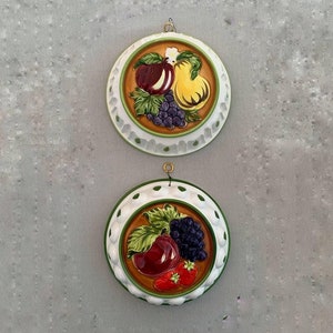 May include: Two white ceramic cake moulds with green and brown trim. The moulds feature a raised design of fruit, including apples, pears, grapes, and strawberries.