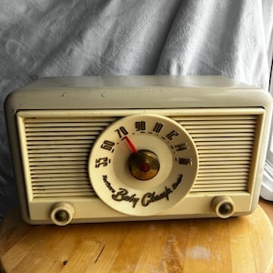 May include: Cream-coloured vintage radio with a circular dial and the words "Northern Baby Champ" printed on the face. The radio has a horizontal speaker grill and two knobs. The dial needle points to the number 70.