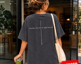 Può includere: Una t-shirt a maniche corte grigio antracite con una croce bianca verticale stampata sul retro. Il testo "THE WAY, THE TRUTH, AND THE LIFE" è stampato orizzontalmente sulla croce, con "JOHN 14:6" scritto verticalmente sotto.