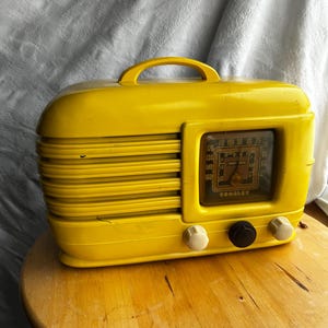 Op de afbeelding: Een vintage gele Crosley-radio met een handvat, wijzerplaat en knoppen. De radio heeft een rechthoekige vorm met horizontale lijnen aan de linkerkant en een helder wijzerplaatvenster. De radio staat op een houten oppervlak.