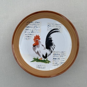 Puede incluir: Un plato de cerámica marrón y blanco con un diseño de gallo. El plato tiene un borde marrón y un centro blanco con una ilustración de gallo blanco y negro. El gallo está de pie sobre un parche de hierba verde. El plato tiene texto escrito a mano en tinta negra alrededor del gallo.