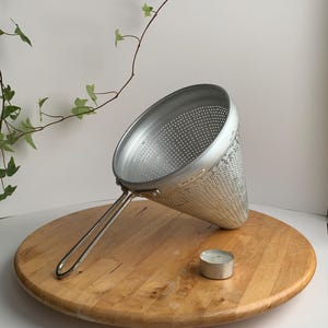 May include: A silver cone-shaped strainer with a handle, sitting on a wooden surface. A small, silver candle is also on the surface.