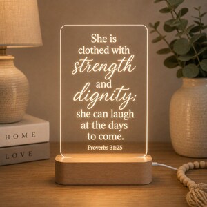 May include: A rectangular acrylic light plaque with a wooden base, featuring the engraved text: She is clothed with strength and dignity; she can laugh at the days to come. Proverbs 31:25. The sign emits a warm glow, illuminating the white lettering.