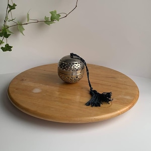 Pode incluir: Um infusor de chá prateado com uma borla preta está sobre uma bandeja grande, redonda e de madeira castanha clara. O infusor tem um design perfurado. Ramos de hera verde estão no fundo.