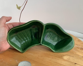 May include: A green ceramic divided dish with a curved shape. The dish is made of glazed ceramic and has a smooth, glossy finish.