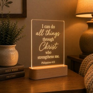 Könnte beinhalten: Eine rechteckige, klare Acryl-Lichtplatte in einem hellen Holzsockel. Die Platte wird mit warmem Licht beleuchtet und zeigt den eingravierten Text: I can do all things through Christ who strengthens me. Philippians 4:13.