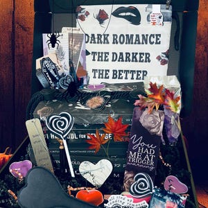 Könnte beinhalten: Eine Geschenkbox mit dem Thema Dark Romance. Die Box enthält Bücher, Lesezeichen, einen herzförmigen Paddel, einen Lolli und andere Gothic-Artikel. Der Text "DARK ROMANCE THE DARKER THE BETTER" ist auf der Box zu sehen.