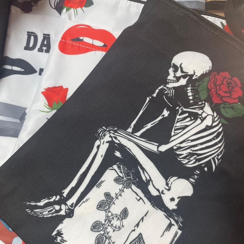 May include: A black tote bag featuring a white graphic of a skeleton sitting on a book with a red rose, partially overlapping a white tote bag printed with red lips, black lips, and red roses, along with the partial text DA.