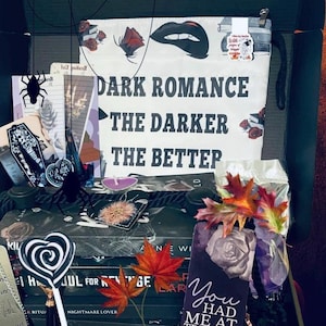 May include: A black gift box containing books and book-themed accessories. Items include a white pouch with the text DARK ROMANCE THE DARKER THE BETTER, a bookmark with the text You HAD ME AT TRIGGER warning, a spider cutout, and artificial leaves.
