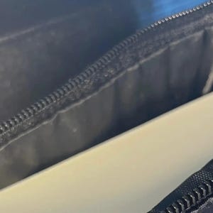May include: A black zippered case with a smooth, matte exterior texture and a black fabric interior lining. The case is partially open, revealing a solid, light-colored, rectangular object tucked inside the main compartment of the storage pouch.