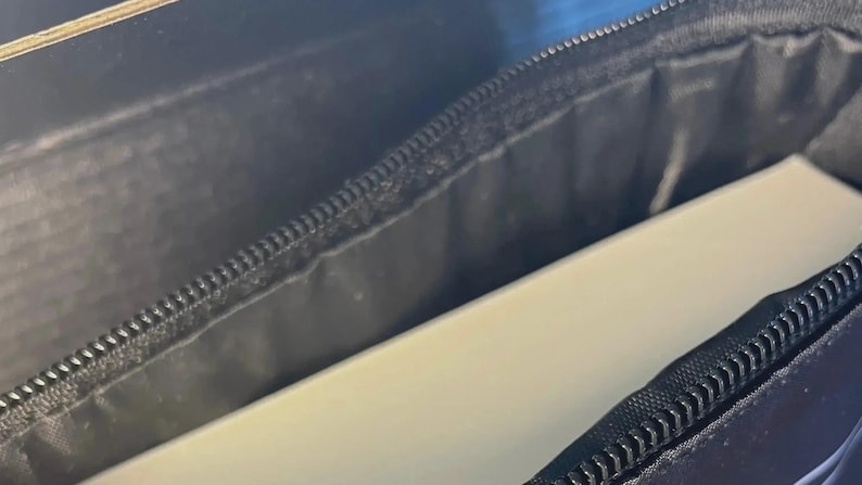 May include: A black zippered case with a smooth, matte exterior texture and a black fabric interior lining. The case is partially open, revealing a solid, light-colored, rectangular object tucked inside the main compartment of the storage pouch.