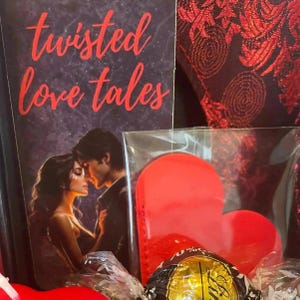 May include: A book titled Twisted Love Tales with a dark cover featuring a couple in an embrace, accompanied by a bright red heart-shaped object in a clear plastic sleeve and a gold-wrapped chocolate candy, all set against a dark red patterned fabric.