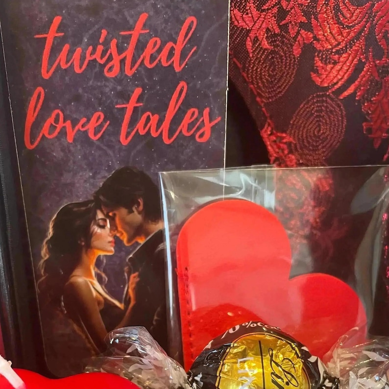May include: A book titled Twisted Love Tales with a dark cover featuring a couple in an embrace, accompanied by a bright red heart-shaped object in a clear plastic sleeve and a gold-wrapped chocolate candy, all set against a dark red patterned fabric.