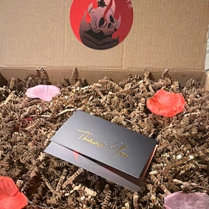 May include: A cardboard shipping box filled with brown crinkle paper, scattered pink and red fabric petals, and a black card with gold foil text that reads Thank You. A circular sticker on the box lid features a skull, candle, and book graphic.