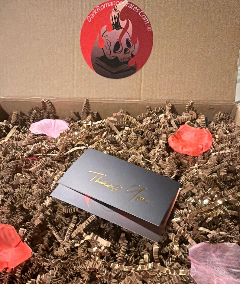 May include: A cardboard shipping box filled with brown crinkle paper, scattered pink and red fabric petals, and a black card with gold foil text that reads Thank You. A circular sticker on the box lid features a skull, candle, and book graphic.