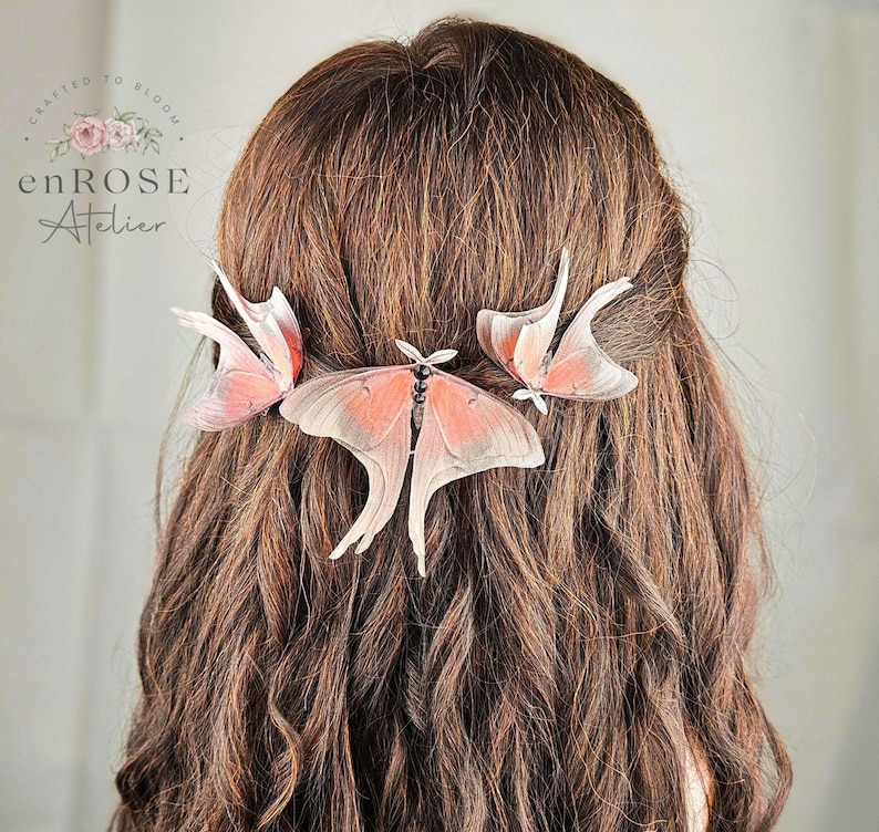 May include: Three delicate pink and white butterfly hair clips, crafted to bloom, are pinned into a woman's long brown hair. The hair clips are made of delicate, translucent material and feature intricate wing details.