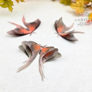 May include: Three delicate, hand-crafted, peach-colored butterfly wings with a gray and white pattern. The wings are arranged on a white surface. enROSE Atelier is printed in black text on the image.