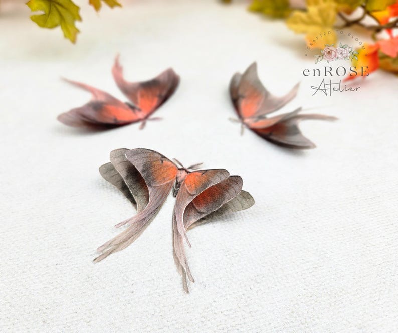 May include: Three delicate, hand-crafted, peach-colored butterfly wings with a gray and white pattern. The wings are arranged on a white surface. enROSE Atelier is printed in black text on the image.
