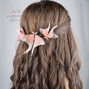 May include: Two delicate pink and white butterfly hair clips are worn in a woman's brown hair. The butterflies are made of fabric and have a realistic appearance.