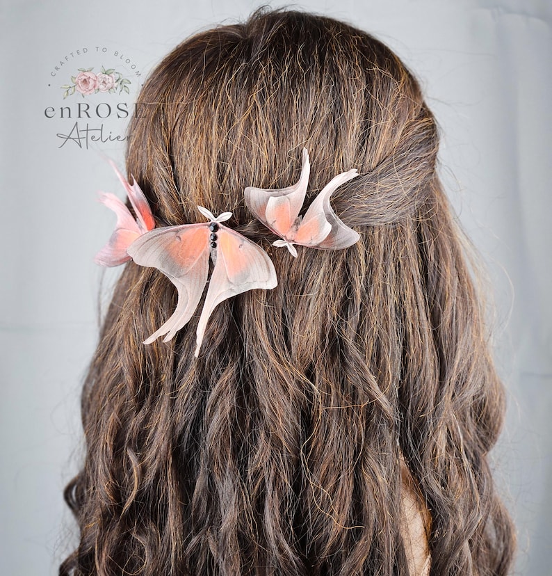 May include: Two delicate pink and white butterfly hair clips are worn in a woman's brown hair. The butterflies are made of fabric and have a realistic appearance.