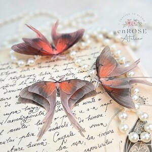 May include: Three delicate, hand-crafted butterfly wings made of sheer fabric with intricate details. The wings are arranged on a vintage-looking page with a pearl necklace draped over it. The text "CRAFTED TO BLOOM enROSE Atelier" is visible in the top right corner.
