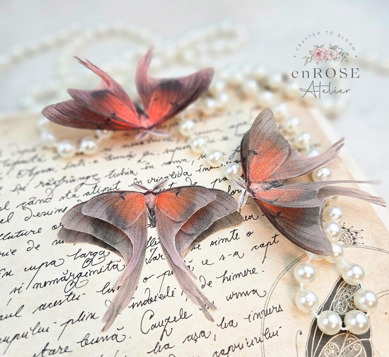May include: Three delicate, hand-crafted butterfly wings made of sheer fabric with intricate details. The wings are arranged on a vintage-looking page with a pearl necklace draped over it. The text "CRAFTED TO BLOOM enROSE Atelier" is visible in the top right corner.