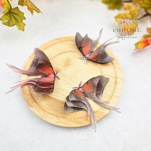 May include: Three delicate, hand-painted butterfly wings in shades of pink and brown, arranged on a wooden surface. The wings are layered and have a realistic appearance.