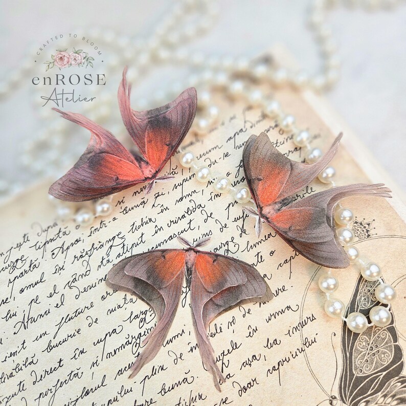May include: Three delicate, translucent butterfly wings in shades of pink and brown are arranged on a vintage, cream-colored page with cursive writing. The wings are layered on top of each other, creating a beautiful and ethereal display. A strand of white pearls is draped across the page, adding a touch of elegance.