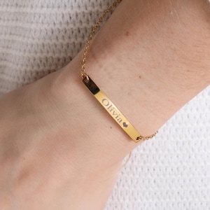 Puede incluir: Una pulsera dorada con una barra grabada con el nombre "Olivia" y un símbolo de corazón. La pulsera está unida a una fina cadena. La pulsera se lleva en la muñeca.