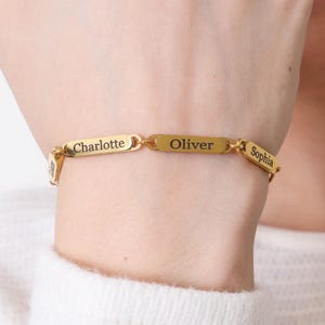 May include: Gold-toned bracelet with the names "Charlotte", "Oliver", and "Sophia" engraved on rectangular links. The bracelet is worn on a wrist, showcasing the personalized jewelry.