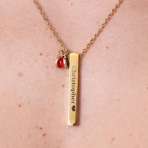 May include: Gold-colored necklace with a rectangular pendant engraved with the name "Christopher" and a heart symbol. The necklace features a chain and two small red gemstone charms.