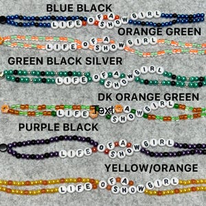 May include: A collection of beaded bracelets in various color combinations, including blue and black, orange and green, and purple and black. Each bracelet spells out "LIFE OF A SHOW GIRL" with white letter beads.
