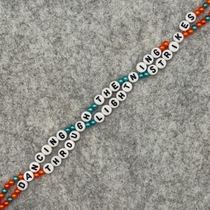 Two strands of beaded bracelets with the words "DANCING THROUGH THE LIGHTNING STRIKES" spelled out in white letter beads. The beads are strung on orange and teal colored string, against a gray background.
