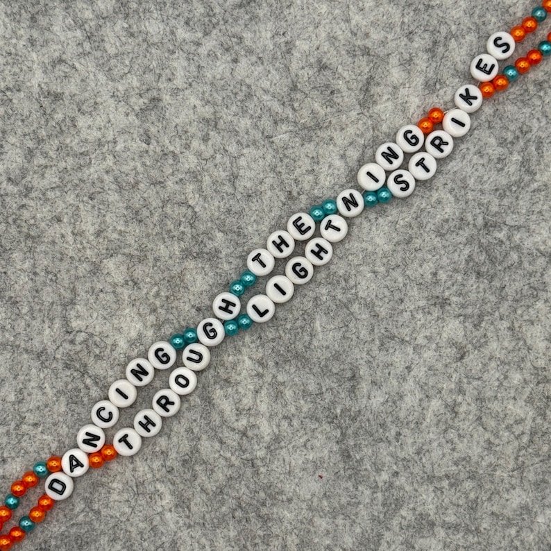Two strands of beaded bracelets with the words "DANCING THROUGH THE LIGHTNING STRIKES" spelled out in white letter beads. The beads are strung on orange and teal colored string, against a gray background.