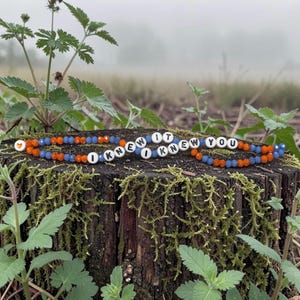 May include: A beaded bracelet featuring alternating orange and blue faceted beads, white letter beads spelling out I KNEW IT I KNEW YOU, and a single orange heart bead. The bracelet is arranged in a circular shape on a mossy tree stump.