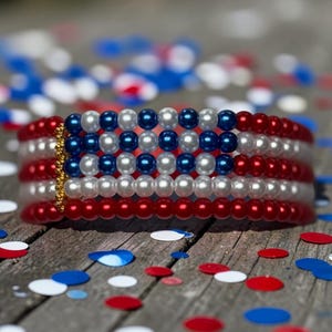 Op de afbeelding: Een patriottische armband met rode, witte en blauwe kralen die de Amerikaanse vlag voorstellen. De ronde kralen zijn klein, met een gouden sluiting. Rood, wit en blauw confetti ligt verspreid op een houten oppervlak.