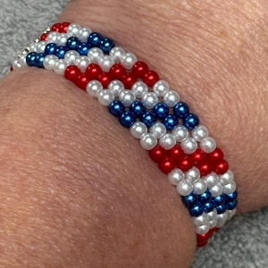 A beaded bracelet made up of 4 rows of red, white, and blue beads in the pattern of 2 red, 2 white, 2 blue. The bracelet is made of small, round beads, and the colors are arranged in alternating rows. The bracelet is worn on a wrist.