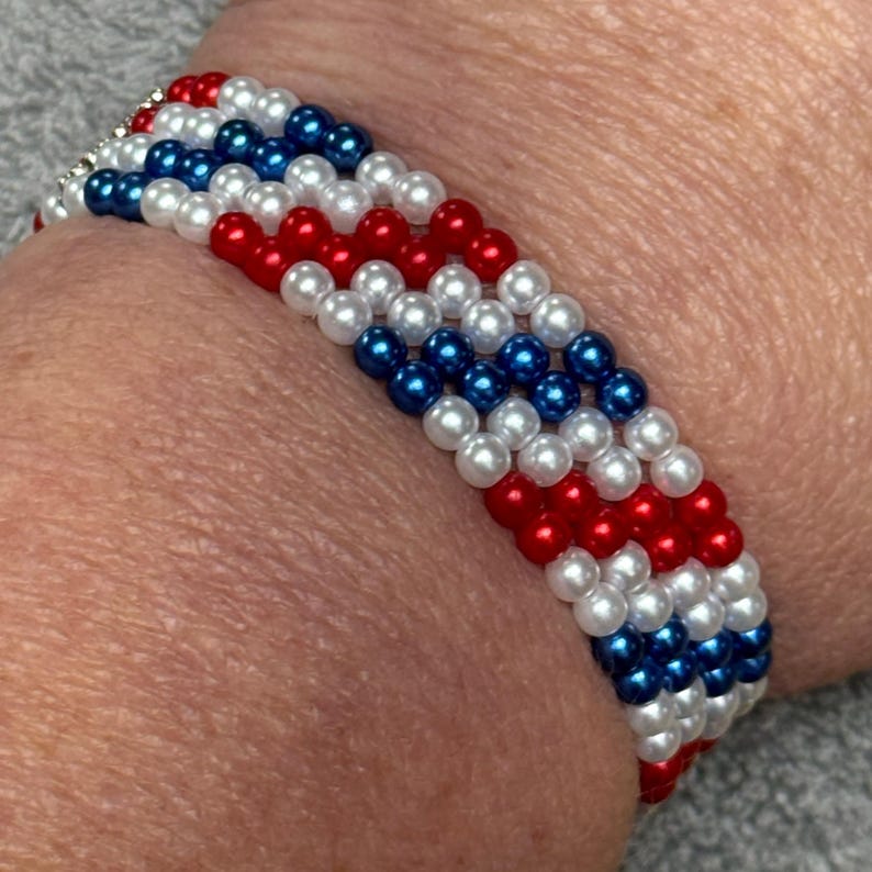 A beaded bracelet made up of 4 rows of red, white, and blue beads in the pattern of 2 red, 2 white, 2 blue. The bracelet is made of small, round beads, and the colors are arranged in alternating rows. The bracelet is worn on a wrist.