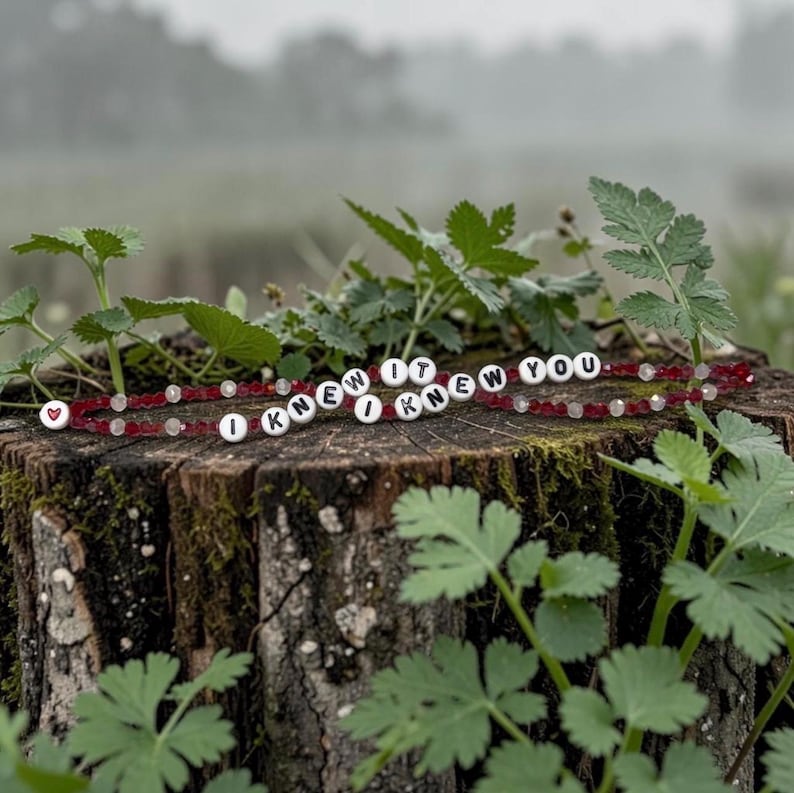 May include: A beaded bracelet featuring alternating red and clear faceted beads with white circular letter beads spelling out the phrase I KNEW IT I KNEW YOU and a single white bead with a small red heart symbol at the beginning of the strand.