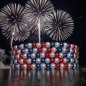 Op de afbeelding: Een decoratief, cilindrisch object gemaakt van rode, witte en blauwe kralen, dat lijkt op een armband of een feestelijke display. Vuurwerk barst uit in de nachtelijke hemel boven het object, dat weerspiegeld wordt in het water. De kralen zijn in afwisselende rijen van kleur gerangschikt.