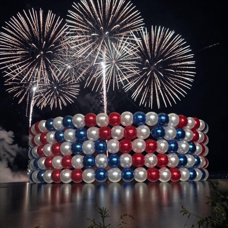 Op de afbeelding: Een decoratief, cilindrisch object gemaakt van rode, witte en blauwe kralen, dat lijkt op een armband of een feestelijke display. Vuurwerk barst uit in de nachtelijke hemel boven het object, dat weerspiegeld wordt in het water. De kralen zijn in afwisselende rijen van kleur gerangschikt.