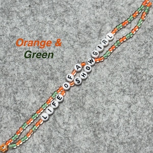 May include: A beaded lanyard with the words "LIFE OF A SHOW GIRL" spelled out in white letter beads. The lanyard features orange and green beads, with a small decorative charm at the end. The words "Orange & Green" are written in the upper left corner.