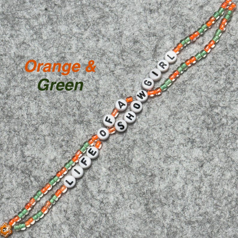 May include: A beaded lanyard with the words "LIFE OF A SHOW GIRL" spelled out in white letter beads. The lanyard features orange and green beads, with a small decorative charm at the end. The words "Orange & Green" are written in the upper left corner.