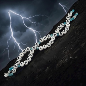 May include: A beaded bracelet with white letter beads spelling "DANCING THROUGH THE LIGHTNING STRIKES". The bracelet has light blue beads and a clear flower-shaped bead at the end. The background shows a dark, textured surface and a lightning storm.