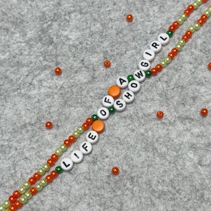 Bracelet made of 2 intertwining strings of 4mm dia light green and orange beads with white beads with black letters that say "LIFE OF A SHOWGIRL"