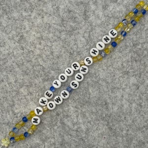 May include: A beaded lanyard with the words "MAKE YOUR OWN SUN SHINE" spelled out in white letter beads. The lanyard features yellow and blue beads, and a small flower charm at the end.